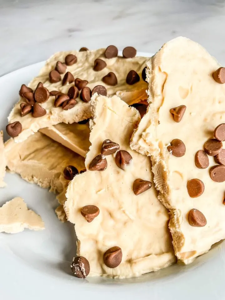 Chocolate chip cookie cake with creamy frosting on white plate, sliced to show layers.