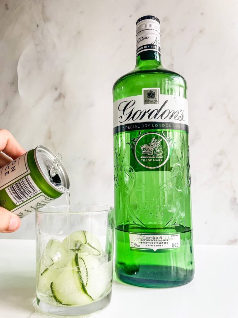 Cucumber ice cubes in glass with tonic water being poured on top next to bottle of gin